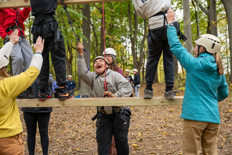 The image shows a group of people on a high ropes course. Some are on the wooden beams, while others are on the ground. They are wearing safety gear, including helmets and harnesses. The people on the course appear to be enjoying themselves, with some raising their arms in the air.